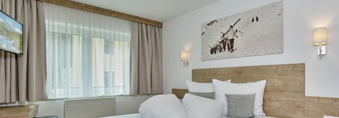 A modern bedroom with a large bed and wooden details. A picture of snow and mountains hangs on the wall. - Gästehaus Bergkristall - Sölden