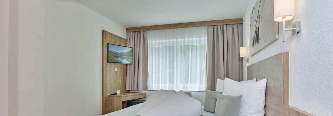 A modern bedroom with a large bed and bright, cheerful colors. There are windows with curtains and a small TV corner. - Gästehaus Bergkristall - Sölden