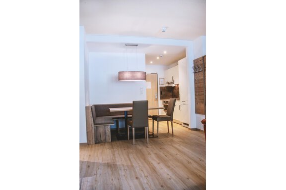 A modern dining room with a wooden table and chairs. There is a bright ceiling light and an open kitchen in the background. - Gästehaus Europa - Sölden