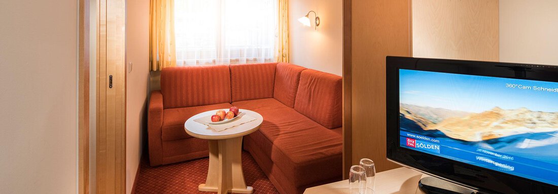 A cozy hotel room with a red sofa, a table, and a television. The light filters through a window, creating an inviting atmosphere. - Gästehaus Europa - Sölden