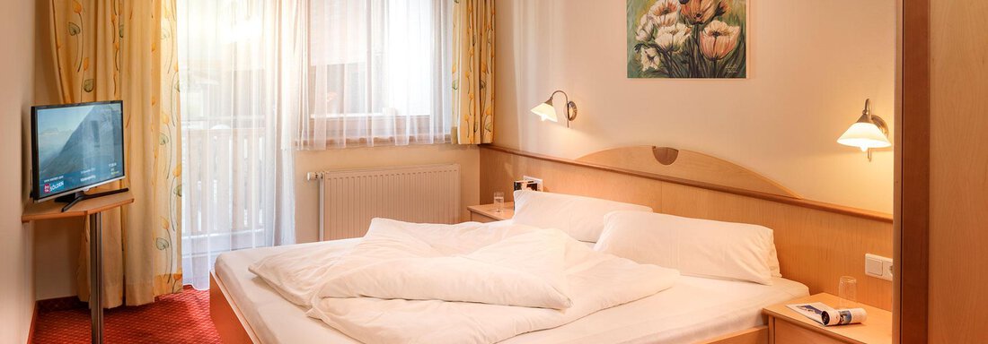 A cozy hotel room with a double bed and two nightstands. A television stands in the corner, and the window lets in plenty of daylight. - Gästehaus Europa - Sölden