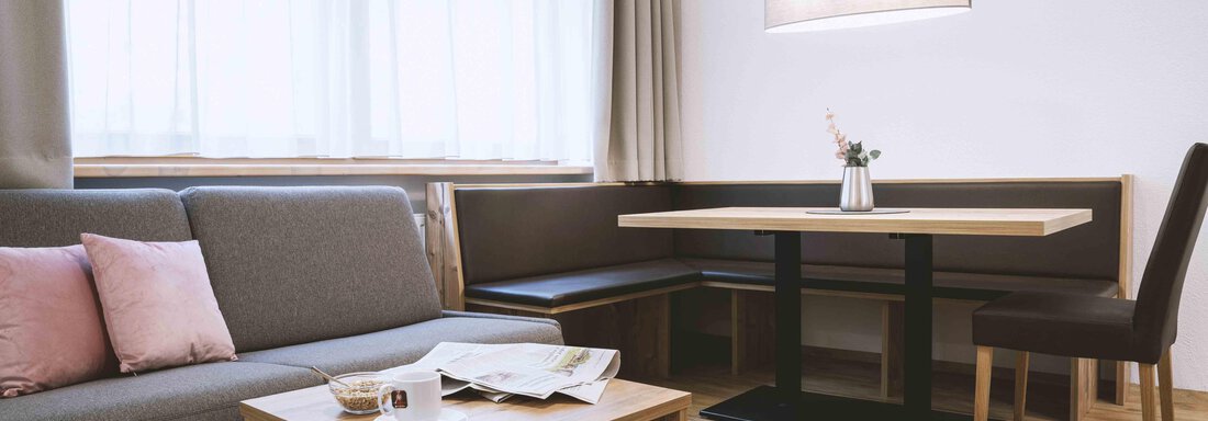 A cozy living space with a gray sofa, a wooden table, and a dining set. Large windows provide plenty of daylight. - Gästehaus Europa - Sölden
