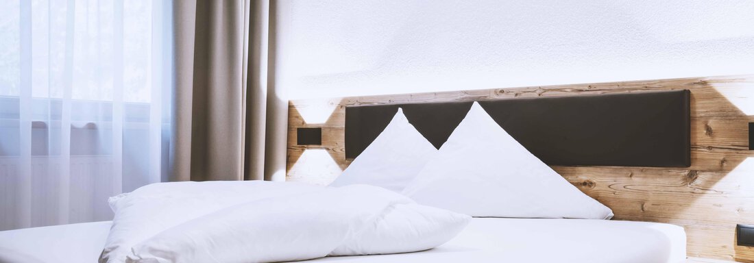 A modern bedroom with a wooden frame bed and two pillows. Bright curtains hang in front of the window, creating a bright atmosphere. - Gästehaus Europa - Sölden