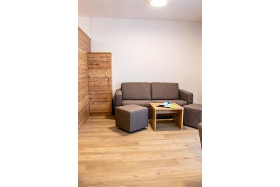 A modern living room with a gray sofa, a wooden table, and cozy ottomans. The walls are light and the floor is wooden. - Gästehaus Europa - Sölden