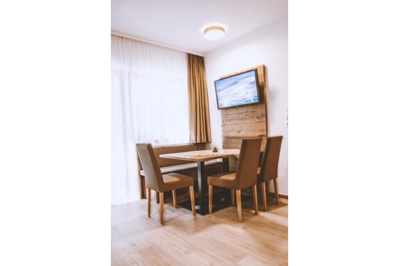 A cozy dining area with a wooden table and four chairs. In the background, a television is mounted on the wall, and large windows let in plenty of light. - Gästehaus Europa - Sölden