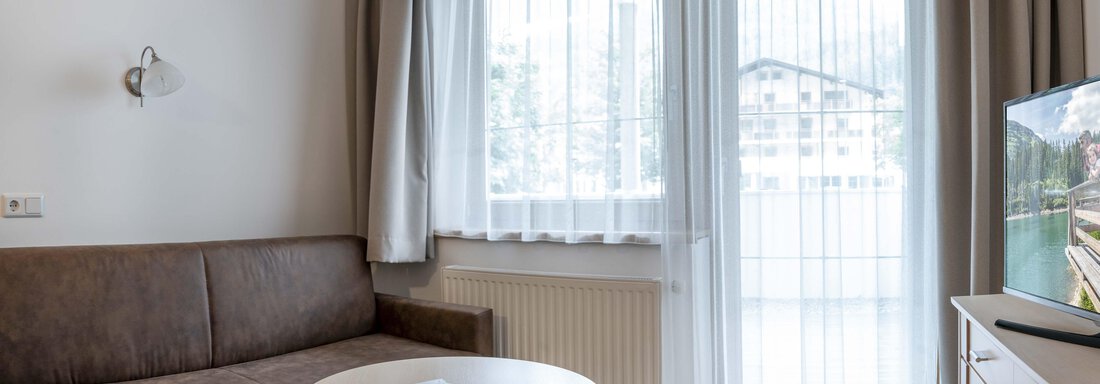 A cozy living space with a brown sofa and a round table. Large windows with curtains let in plenty of light. - Gästehaus Europa - Sölden
