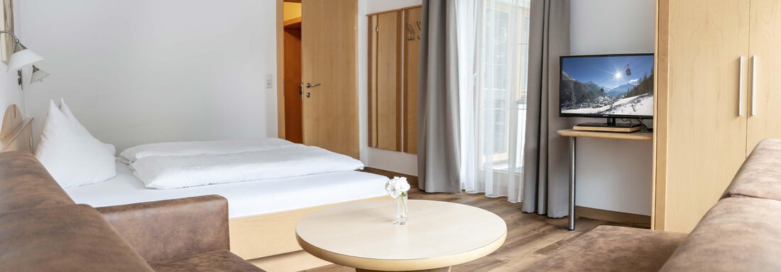 A modern room with a double bed and a cozy seating area. The room has a television and large windows that let in a lot of light. - Gästehaus Europa - Sölden