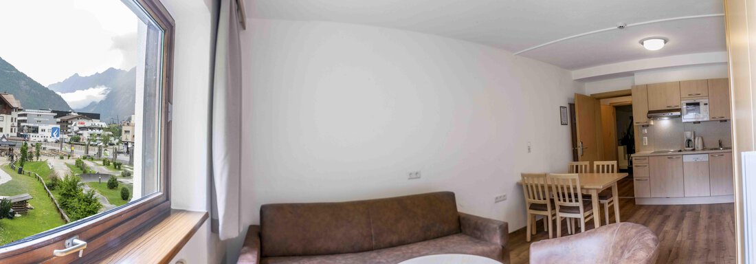 A cozy living space with a window overlooking mountains and a small park. Inside, there is a sofa, a table, and a kitchenette. - Gästehaus Europa - Sölden