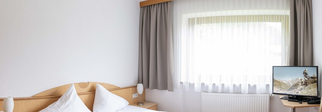 A cozy hotel room with a double bed and a TV. The room is bright and has large windows with curtains. - Gästehaus Europa - Sölden
