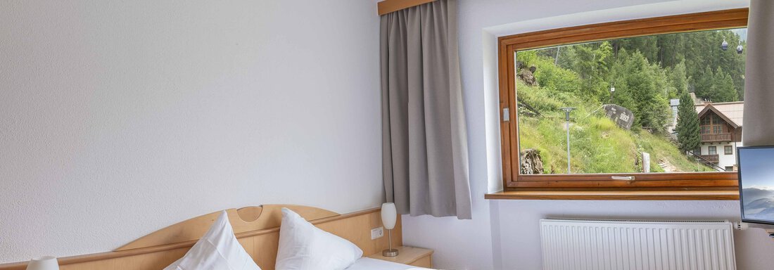 A bright bedroom with a double bed and two pillows. The view from the window shows a green landscape with trees. - Gästehaus Europa - Sölden