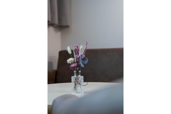 A beautiful bouquet of flowers in a clear glass vase is on a table. In the background, a cozy sofa can be seen. - Gästehaus Europa - Sölden