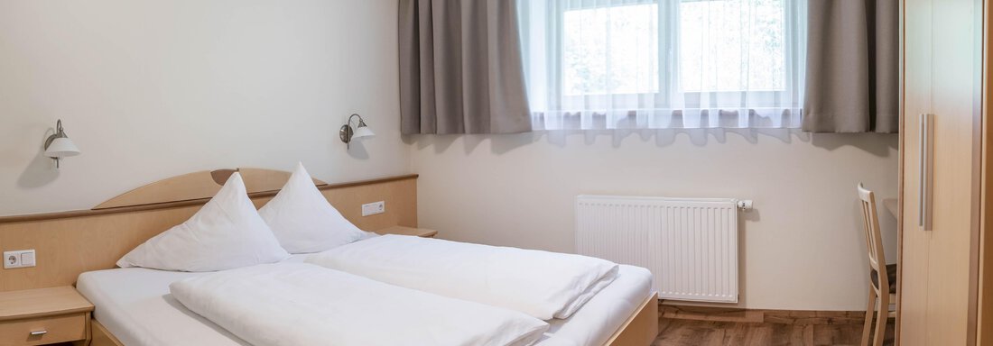 A bright bedroom with a large double bed and two pillows. The room has furniture made of light wood and a large window with curtains. - Gästehaus Europa - Sölden