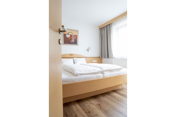 A bright bedroom with a large bed and a wooden floor. A picture hangs on the wall, and the window lets in a lot of daylight. - Gästehaus Europa - Sölden