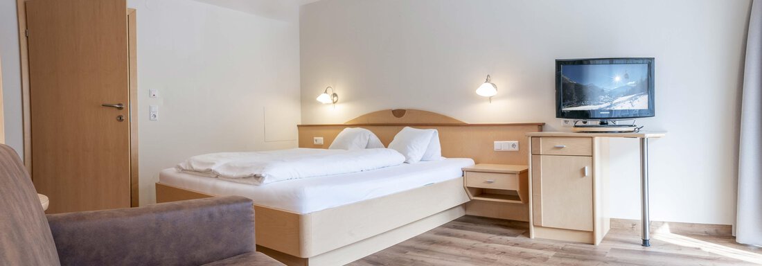 A bright and modern hotel room with a double bed, a television, and a comfortable sofa. The room has wooden flooring and a friendly atmosphere. - Gästehaus Europa - Sölden