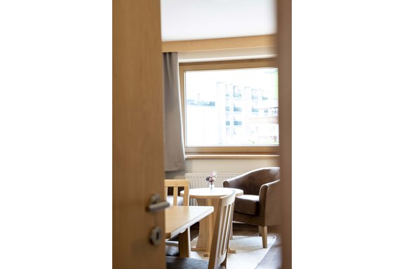 A bright room with a table and chairs as well as a cozy armchair. Through the window, you can see the city. - Gästehaus Europa - Sölden