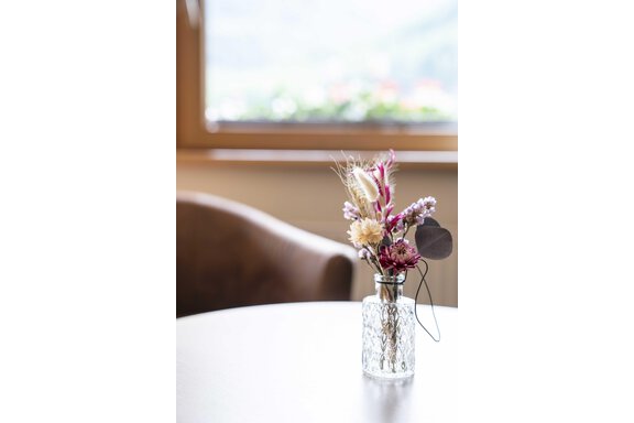 A small vase with colorful dried flowers is on a table. In the background, a window can be seen with a view of the landscape. - Gästehaus Europa - Sölden