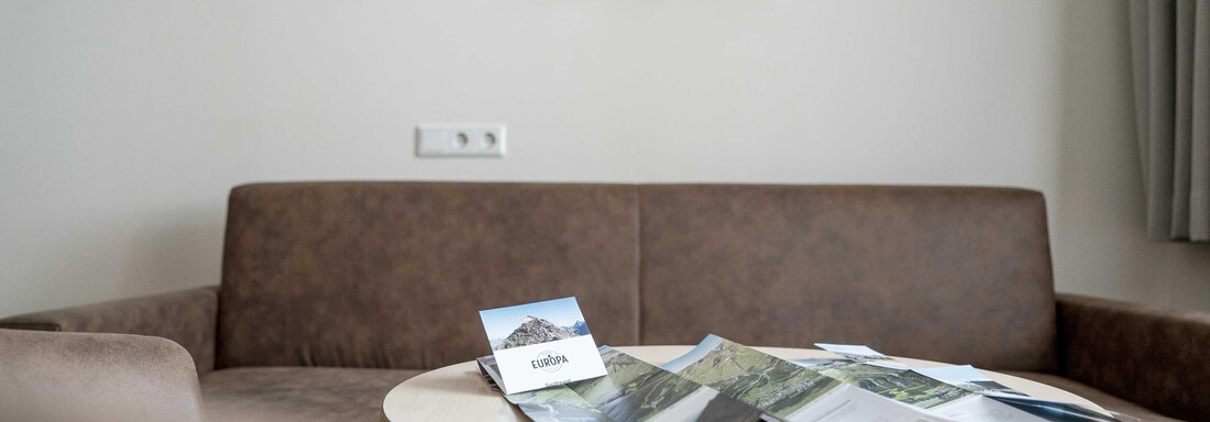 A cozy sofa with a round table on which some brochures are lying. Light sources are hanging on the wall, and the room feels bright and inviting. - Gästehaus Europa - Sölden