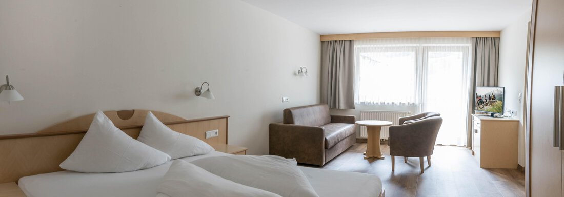 A modern hotel room with two beds and a cozy seating area. Large windows let in plenty of daylight and offer a view outside. - Gästehaus Europa - Sölden