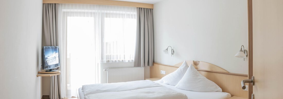 A cozy hotel room with a double bed and a flat-screen TV. Large windows provide plenty of daylight and access to the balcony. - Gästehaus Europa - Sölden