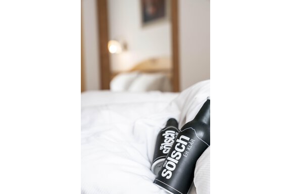 Two bottles with the inscription "söisch" are lying on a bed with white bedding. In the background, there is a mirror that emphasizes the cozy atmosphere of the room. - Gästehaus Europa - Sölden