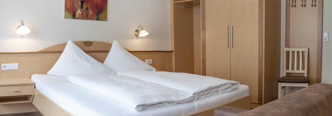 A bright room with a double bed and modern furniture. A colorful painting hangs on the wall, and there is a wardrobe as well as a chair. - Gästehaus Europa - Sölden
