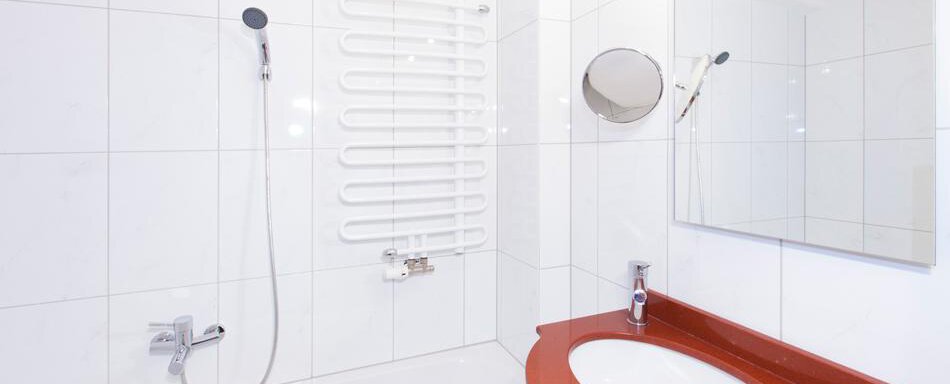 A modern bathroom with white tiles and a shower. The fixtures include a sink with a red edge and a mirror. - Gästehaus Kneisl - Sölden