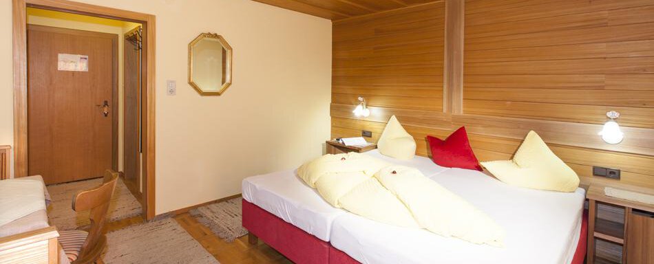 A cozy hotel room with wooden decor. There is a large bed with pillows and a table with a chair. - Gästehaus Kneisl - Sölden
