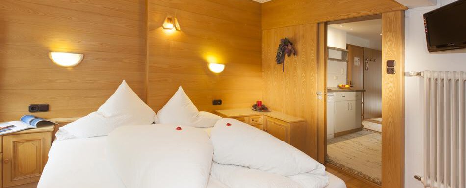 A cozy bedroom with wooden walls and a large bed. The room has a bright atmosphere and leads to a small kitchen. - Gästehaus Kneisl - Sölden