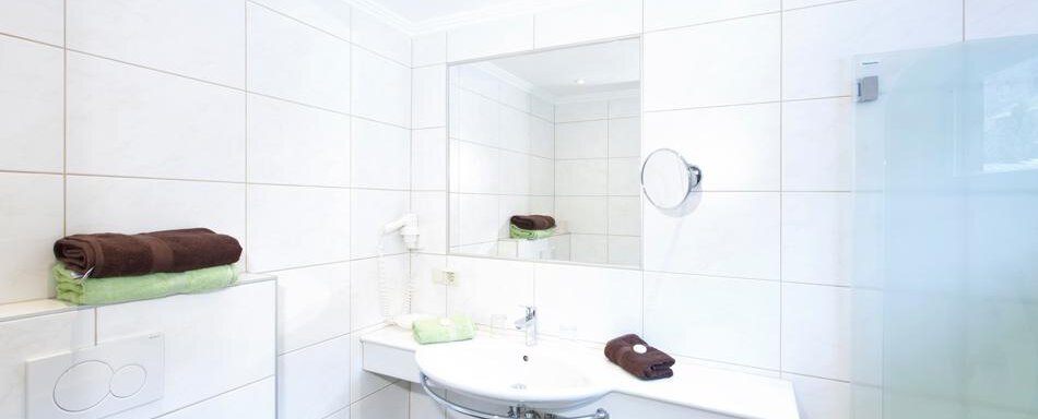 A modern bathroom with white tiles and an elegant sink. Towels in brown and green are neatly arranged. - Gästehaus Kneisl - Sölden