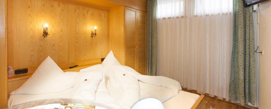 Gaestehaus-Kneisl-Apart-Komfort1 A cozy bedroom with wooden walls and a large bed. The windows are equipped with curtains and there is a small wall console with a television. - Gästehaus Kneisl - Sölden