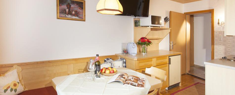 A cozy dining area with a table, chairs, and a small kitchenette. Pictures hang on the wall, and fruits and a bottle of water are on the table. - Gästehaus Kneisl - Sölden