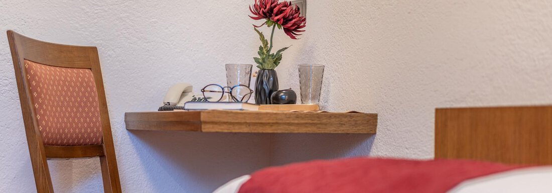 A cozy room with a wooden chair and a small table. On the table, there is a flower and some personal items. - Gästehaus Maier - Sölden