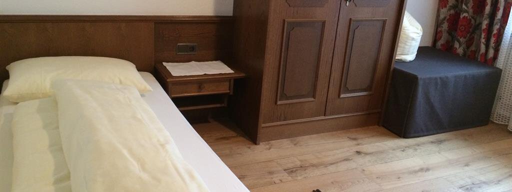 Einzelzimmer A simple bedroom with a wooden bed and a box. The room has a bright wooden floor and a curtain with a floral pattern. - Gästehaus Martin - Sölden