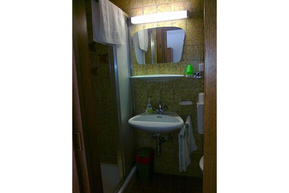A small bathroom with a shower, a sink, and a mirror. The walls are decorated with brown tiles, and there is a trash can and towels. - Gästehaus Martin - Sölden