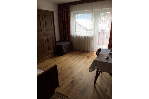 A simple room with wooden flooring and large windows. A table and a chair are available, as well as a closet and a small stool. - Gästehaus Martin - Sölden