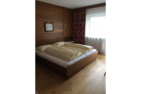 A simple bedroom with a double bed and wooden paneling. Curtains are hanging in front of the window, and the floor is made of wood. - Gästehaus Martin - Sölden