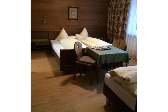 Dreibettzimmer A cozy room with two beds and a small table. The walls are made of wood and there is a curtain with a floral pattern. - Gästehaus Martin - Sölden