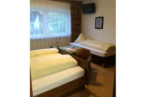 A simple room with two beds, a television, and a window. The walls are adorned with curtains, and there is a small table. - Gästehaus Martin - Sölden