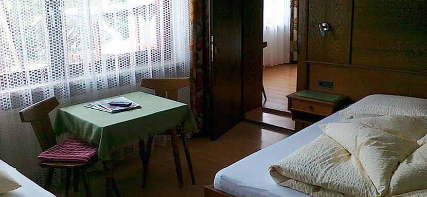 A simple bedroom with two beds and a table. The room has large windows with curtains and a warm, inviting wooden style. - Gästehaus Martin - Sölden
