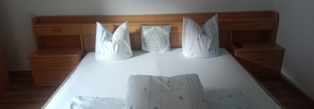 A cozy bedroom with a big bed and white pillows. The walls are light and there are wooden shelves next to the bed. - Gästehaus Sternberger - Sölden