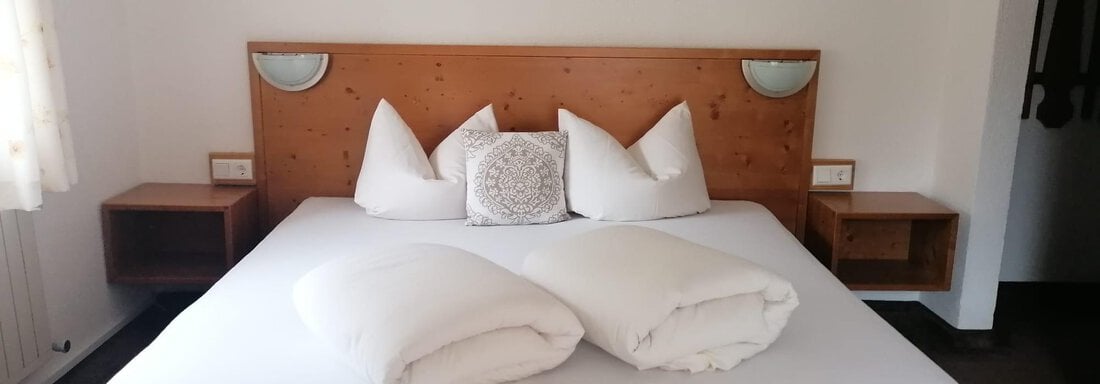 A cozy bedroom with a large bed and small nightstands. White pillows and blankets are on the bed. - Gästehaus Sternberger - Sölden