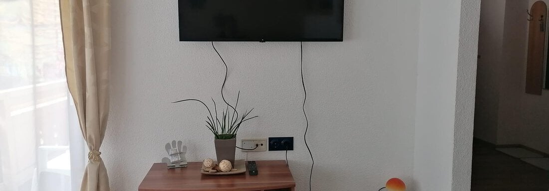 A living area with a TV on the wall. Below it is a wooden shelf with some decorative objects and a plant. - Gästehaus Sternberger - Sölden