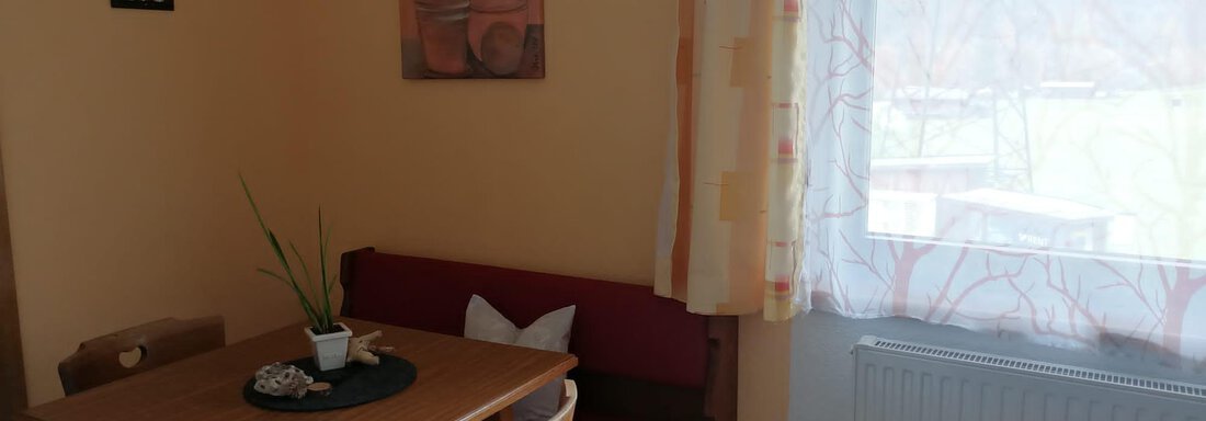 A cozy dining area with a wooden table and red benches. The light shines through a window with a sheer curtain. - Gästehaus Sternberger - Sölden