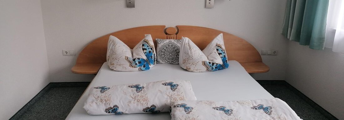 A simple bedroom with a wooden bed and decorative cushions. The walls are painted light and there are some pictures on the wall. - Gästehaus Sternberger - Sölden