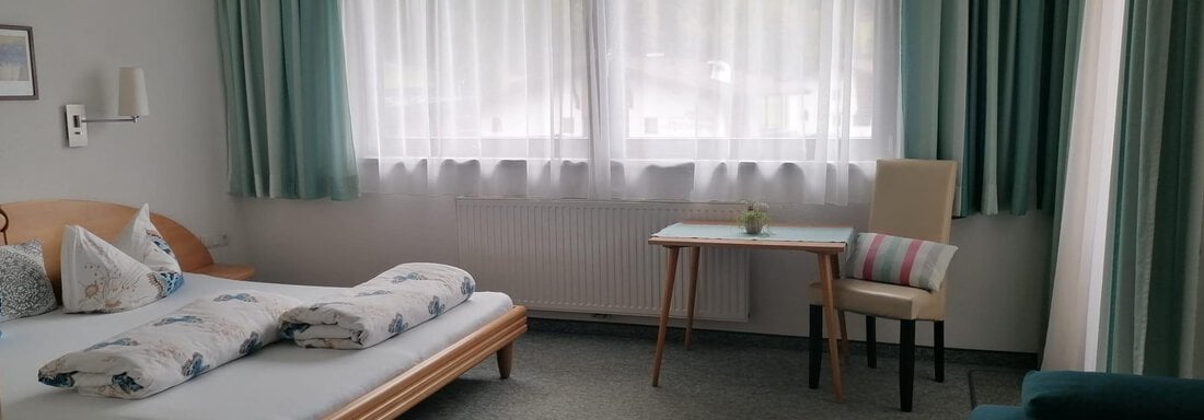 A simple bedroom with a double bed, a table, and a chair. The large windows allow plenty of light in, and the room is bright and welcoming. - Gästehaus Sternberger - Sölden