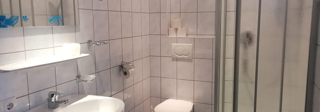 A modern bathroom with white tiles and blue accents. It features a shower, a sink, and a toilet. - Gästehaus Sternberger - Sölden