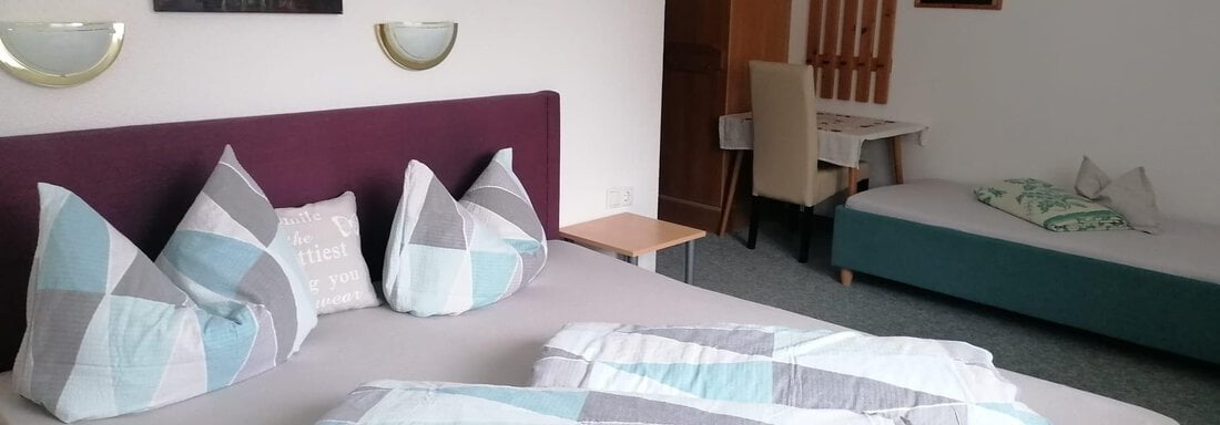 A cozy bedroom with a large bed and decorative pillows. In the background, there is a table and another sleeping area. - Gästehaus Sternberger - Sölden