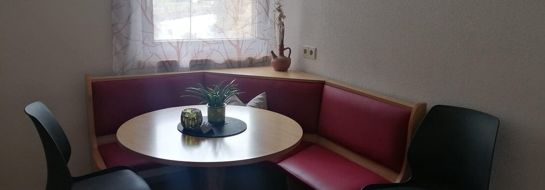 A cozy dining area with a round table and a red bench. Next to it are two black chairs and a window with a view outside. - Gästehaus Sternberger - Sölden