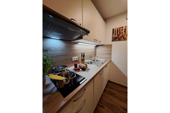 A modern kitchen with wooden surfaces and a stove. On the countertop, pots, fresh vegetables, and cooking utensils are visible. - Gästeheim Schmiedhof - Sölden