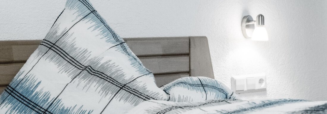 A cozy bed with a checked pillow and a warm blanket. A modern wall lamp is located on the wall. - Gästeheim Schmiedhof - Sölden
